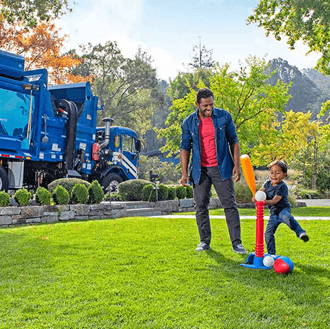 Man and Child Playing in Yard With Garbage Truck Driving By